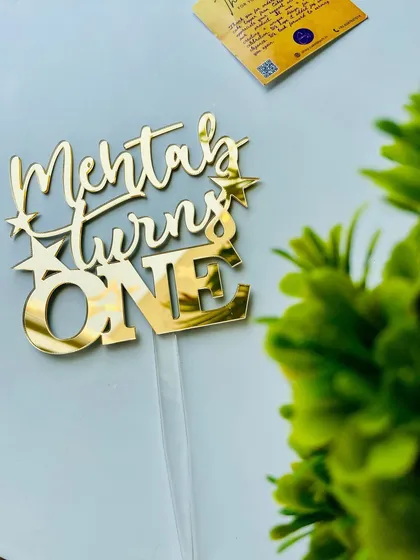 Another view of the "Mehtab turns ONE" topper, this time with a plant in the background, giving it a fresh and natural feel.