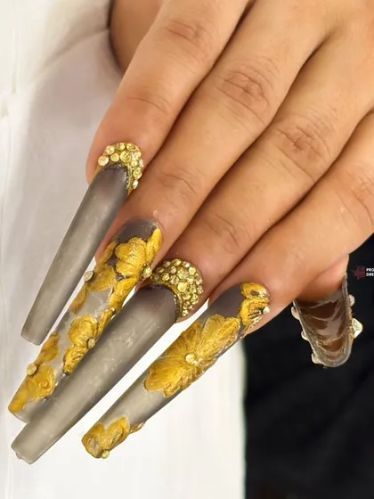 A dramatic and moody design featuring extra-long matte grey nails with raised gold leaf patterns and a cluster of gold crystals at the cuticle.