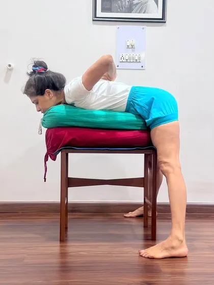 This supported forward bend on a chair with bolsters allows the practitioner to hold the pose without strain. It facilitates the surrender of the mind and body, which is the essence of Isvara pranidhana, or dedication to a higher principle. The props help in achieving physical quietude, which is a prerequisite for mental stillness.