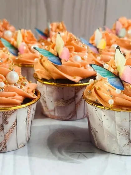 For a larger event, I can prepare bigger batches, like this array of peach butterfly cupcakes. Each one is presented in a beautiful marble-patterned liner with gold trim, enhancing the overall elegant look.