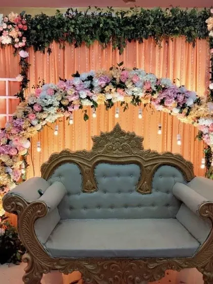A beautiful stage setup for an engagement or roka ceremony. This decor features a floral arch in pastel shades, a classic golden-framed sofa for the couple, and warm lighting to create a romantic ambiance.
