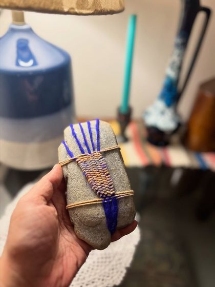 This is the finished woven pebble, held in hand to show its scale and texture. The combination of the hard, smooth stone and the soft, colorful yarn creates a unique tactile object that feels wonderful to hold.