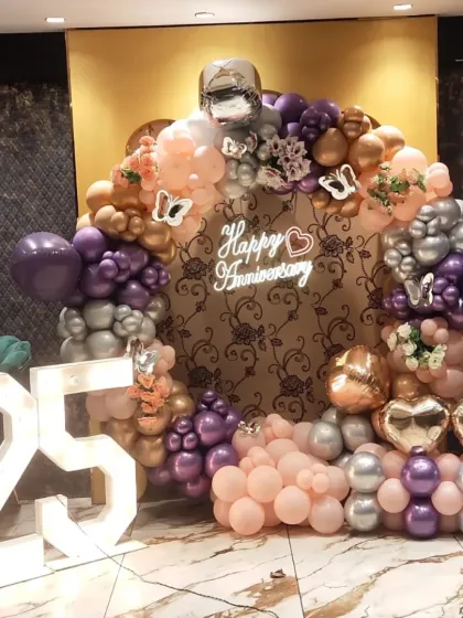 A beautiful 25th-anniversary celebration with a circular balloon arch in purple, peach, and silver. The setup includes a "Happy Anniversary" neon sign and large marquee numbers.