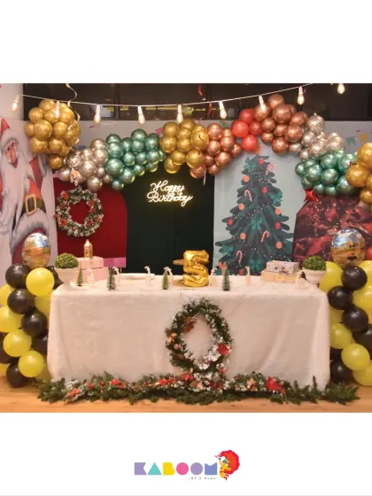 A festive Christmas-themed birthday party, combining holiday cheer with birthday fun. The setup includes a Santa backdrop, wreaths, and a red, green, and gold balloon arch.