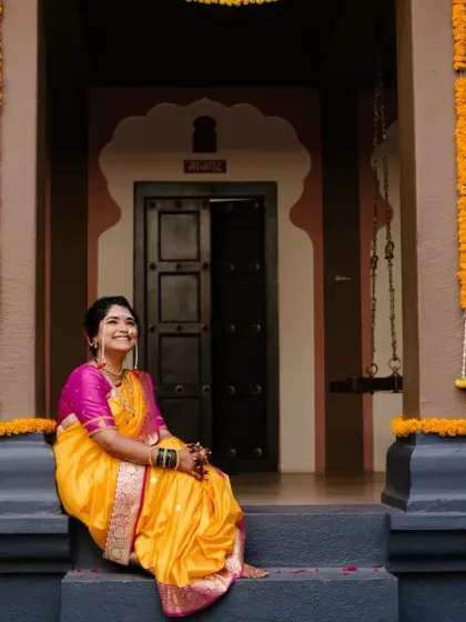 The bride, dressed in a vibrant yellow and pink saree, sits on the steps of a traditional home, looking serene and happy.