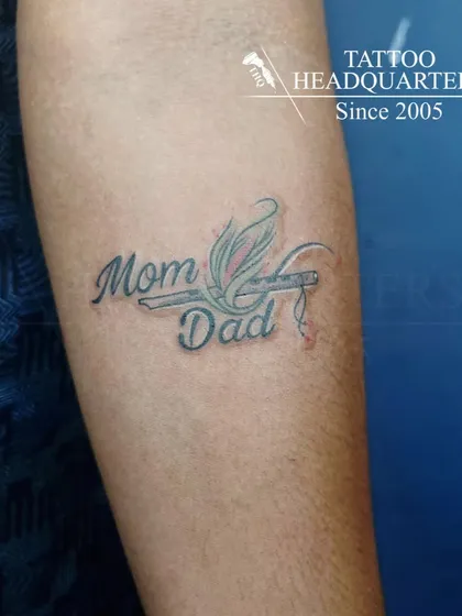 A "Mom Dad" tattoo where the names are connected by a flute and peacock feather, blending family love with spiritual symbols.