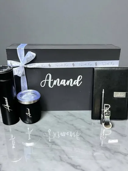 A sleek and modern personalized gift set for 'Anand'. This hamper includes a black thermal flask, a matching tumbler, and a professional diary with a pen, all customized with his name.