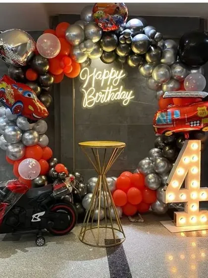 A Cars and fire truck themed birthday party for a 4-year-old. The setup includes a circular balloon arch in red, black, and silver, with a toy bike and marquee number '4'.