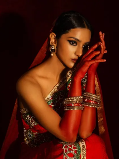 A close-up from the red bridal series, focusing on the henna-like red paint on the hands and the dramatic winged eyeliner. The pose is graceful and draws attention to the details.