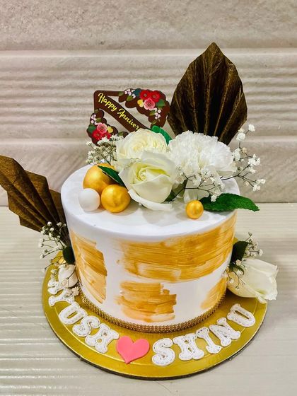 A beautiful cake with a "Light of Gold" theme, featuring a white base with gold brushstrokes and topped with white flowers and dried palm leaves.