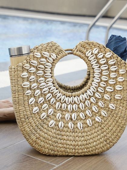 Our shell-adorned woven bag is the perfect poolside or beach companion. Its unique design holds essentials like a bamboo thermos and a towel, blending natural style with practical function for a day of relaxation.