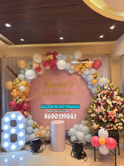 An 8th birthday setup with a large floral arrangement. The pink backdrop and pastel balloon arch are paired with a marquee number '8'.