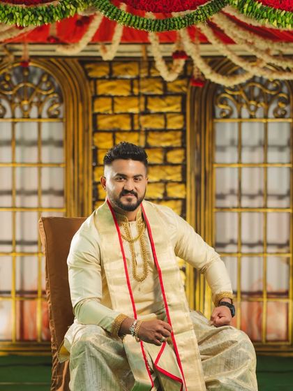 A portrait of the groom in traditional attire, seated within a wedding setup that features a brick-textured backdrop and classic floral garlands.