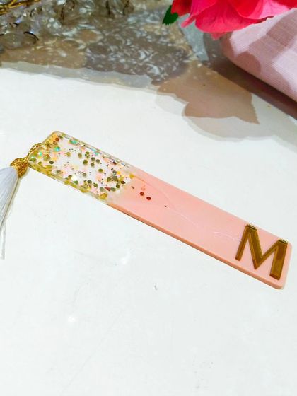 A custom initial bookmark for 'M' in a peachy-pink color with a gold and white glitter top section.