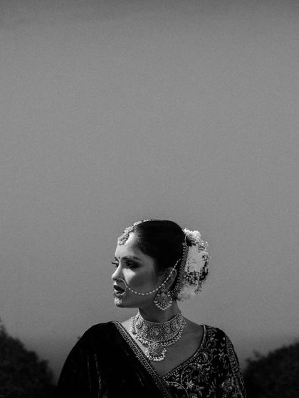 A striking black and white portrait of a bride in profile, her traditional jewelry and hair adorned with flowers standing out against a simple background.