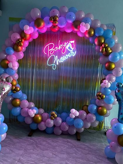 A "He or She" themed baby shower ring in pink, blue, and gold, with a "Baby Shower" neon sign and an iridescent backdrop.