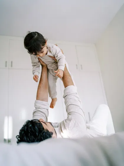 A father lifting his son in the air while lying on the bed. A playful and joyful moment captured during an at-home session.