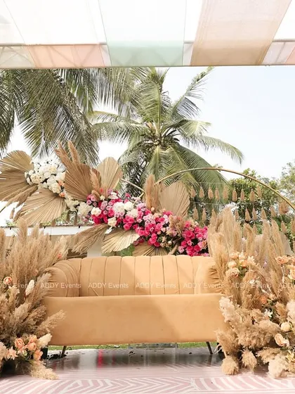 For a sun-drenched Mehndi ceremony, we designed this stunning boho-chic seating area. The arrangement features a comfortable lounge sofa framed by an arc of dried palm leaves, pampas grass, and vibrant pink florals, creating a picture-perfect spot for the couple.