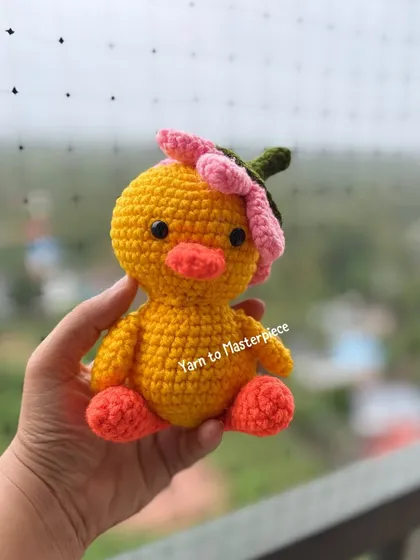A single crochet duck with a pink flower hat, held up to show its cute details. It's small enough to sit on a desk or shelf.