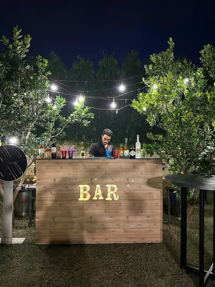 A rustic wooden bar setup for an outdoor Diwali party, nestled between trees and illuminated by string lights.