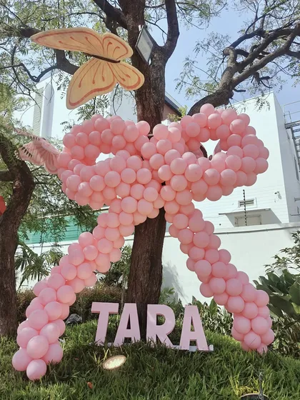 A giant pink balloon bow tied around a tree, personalized with the name "Tara." This is a creative and eye-catching way to add a personalized touch to an outdoor party space.