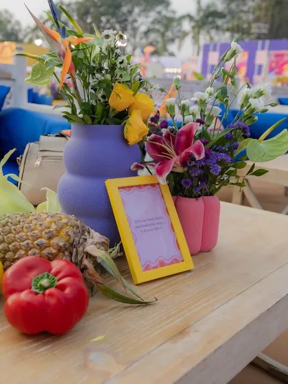 A close-up of a vibrant and playful table setting from a carnival-themed event. Fresh pineapples, bell peppers, and corn are used as unique, sustainable decor elements alongside colorful floral arrangements, demonstrating a fun and unconventional approach to design.