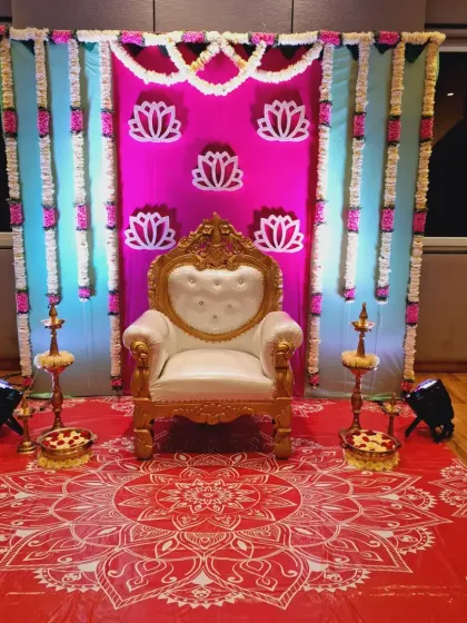 A vibrant Seemantham setup with a red mandala-print floor and a bright pink backdrop with lotus motifs. The lighting and colors make it very festive.