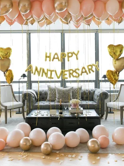 A chic and sophisticated anniversary setup in a modern living room. Rose gold and gold balloons on the ceiling and floor complement the elegant furniture and 'Happy Anniversary' foil letters.