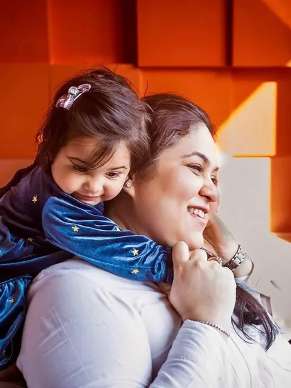 A warm, colorful portrait of a mother and daughter sharing a sweet cuddle. The natural light from the window and the vibrant background create a cozy, happy atmosphere.