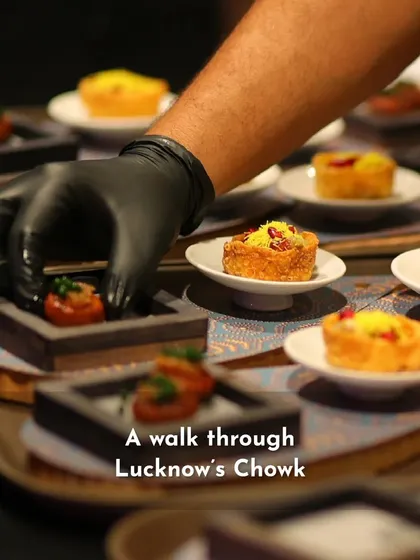 A chef's gloved hand carefully plates an appetizer inspired by the street food of Lucknow's Chowk. This image represents the care taken in presenting regional Indian delicacies with a modern flair.