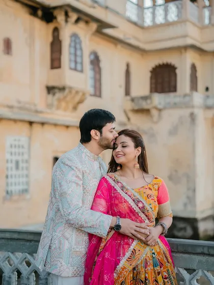A warm and affectionate portrait of the groom kissing the bride's forehead, set against the beautiful architecture of a Udaipur palace.