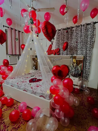 I design romantic room surprises for anniversaries and special moments. This setup features a fairy light canopy over the bed, with red and white balloons, heart-shaped foil balloons, and rose petals to create a truly memorable experience.