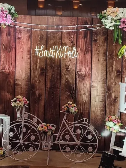 A rustic and romantic photobooth featuring a wooden wall backdrop, a decorative bicycle, and a custom neon hashtag. The pastel floral arrangements add a soft, pretty touch to the overall design.