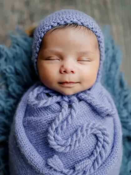 A close-up of the smiling baby from the purple heart basket session. The detailed knit of the bonnet and wrap adds beautiful texture.
