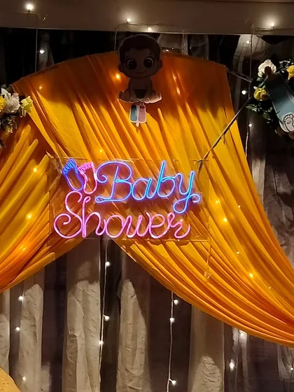 A close-up of the neon sign for a baby shower, with cute baby feet icons, set against a backdrop of yellow fabric and fairy lights.