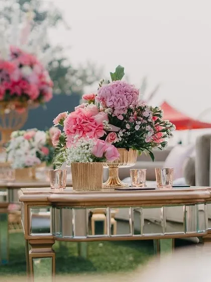 A mirrored coffee table adorned with a beautiful arrangement of pink flowers in a gold vase, reflecting the elegance of the blush-themed wedding.