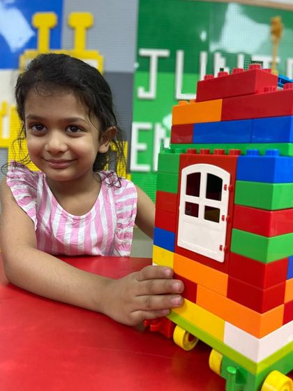 A young girl smiles with her creation, a colorful house on wheels made from large Duplo blocks.