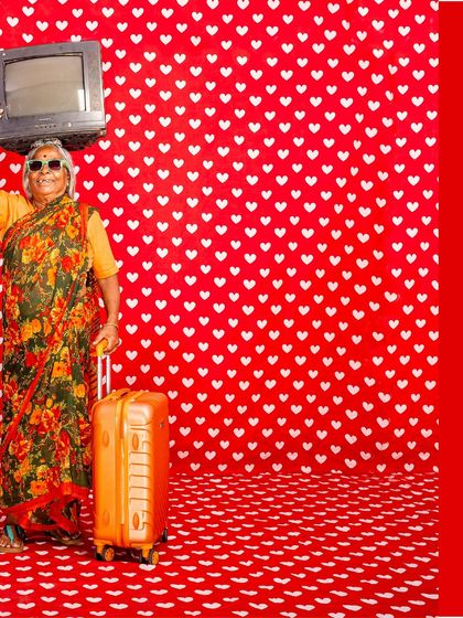Gowramma, the artist's mother, poses with a vintage television and suitcase against a heart-patterned backdrop. This piece from 'No Longer A Memory' is a playful yet poignant commentary on migration, media, and memory.