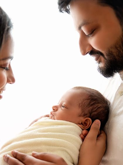 Parents gazing at their newborn, held between them. The backlighting creates a beautiful, ethereal glow around the new family.