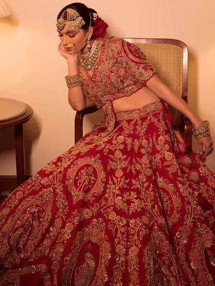 A regal portrait of a bride seated in a chair. The styling of this heavily embroidered red lehenga is designed to look majestic and timeless.