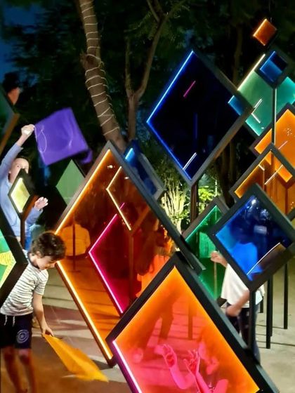 A child explores the colorful, translucent panels of the 'Family Tree' installation. The play of light, color, and reflection creates a magical atmosphere, especially at night.
