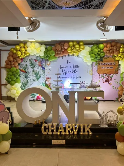 A fun and adventurous jungle safari theme for a first birthday party. The setup includes a lush green and gold balloon arch, a custom backdrop with tropical leaf prints, large "ONE" lettering, and cute safari animal props.