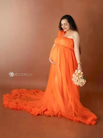 A graceful solo portrait. The one shoulder design and flowing train of the orange gown create a beautiful silhouette, highlighting the baby bump perfectly.