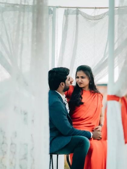 A romantic pre-wedding photo of a couple sharing a look, framed by soft white curtains. The composition creates a dreamy and intimate atmosphere.