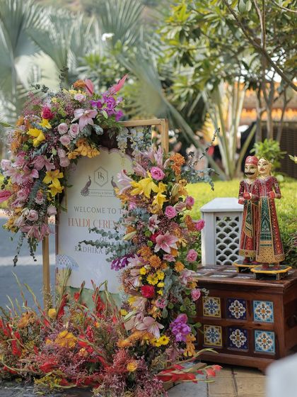 A close-up of the welcome signage, adorned with a lush arch of multi-colored flowers and accompanied by traditional Rajasthani 'kathputli' puppets, adding a touch of local artistry.