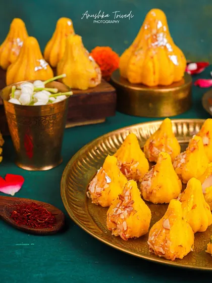 Golden Kesar Modaks, glistening and ready for offering. The deep green background and brass elements create a rich, jewel-toned composition that feels very festive.