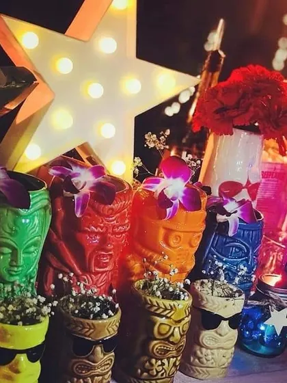 A collection of colorful tiki-style mugs and other retro decor. The glowing star marquee and vibrant flowers add to the fun, nostalgic feel of this 80s-themed cocktail bar.