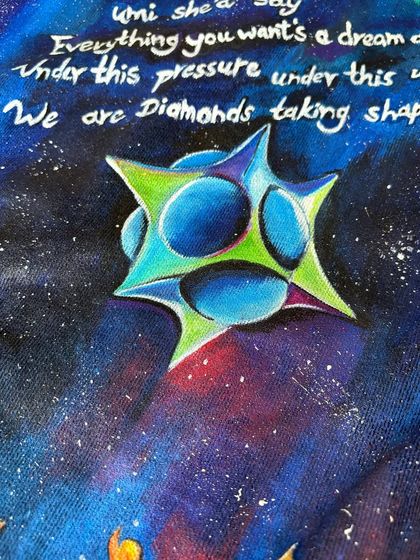 A close-up of the hand-lettered lyrics and the geometric star shape from the "Adventure of a Lifetime" artwork. The white lettering on the dark, starry background creates a powerful visual contrast.