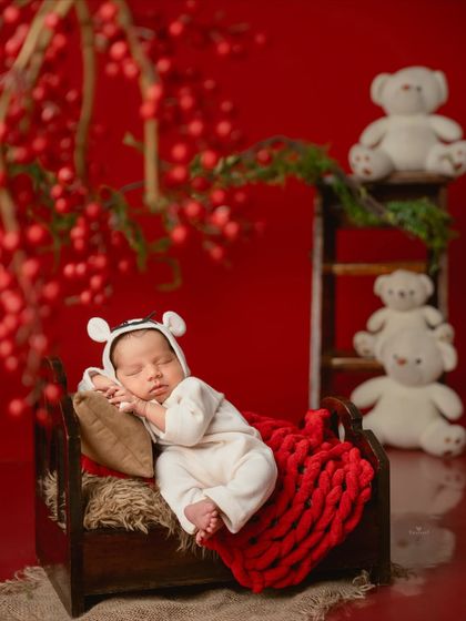 A festive red-themed newborn shoot. The baby, wearing a tiny bear hat, sleeps on a miniature bed with a chunky red knit blanket, perfect for a holiday session.