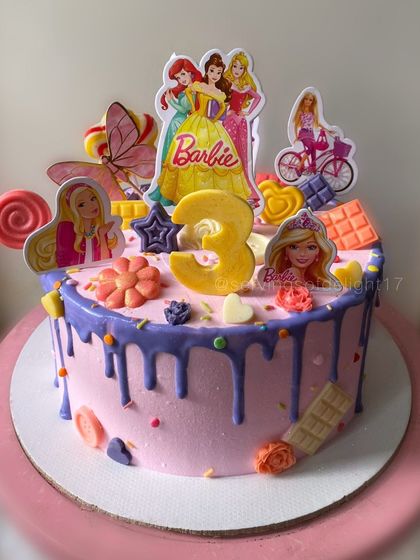 A dream cake for a Barbie fan turning three. This pink drip cake is loaded with Barbie characters, lollipops, and chocolate bars for a fun and festive look.